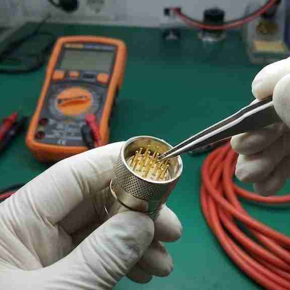 An electronics technician using precision tweezers under a magnifying lamp to inspect bent and recessed gold-plated pins inside a teach pendant cable connector plug to fix intermittent communication errors.