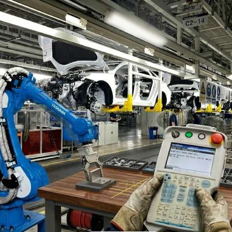 An operator using a Yaskawa DX200 programming pendant in a car factory, illustrating the diagnostic process for robot position loss and encoder feedback instability.